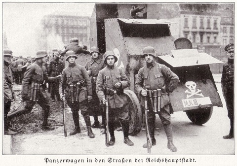 Photo of several men carrying guns. They are dressed in military uniforms and standing in front of a large machine with a skull on it.