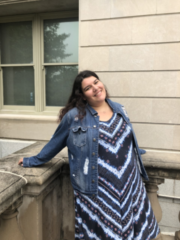 A photo of a woman with long, brown hair smiling at the camera. She is leaning her arms back against a stone balcony. She is wear a denim jacket and a long blue dress with stripes of varying blue shades. 