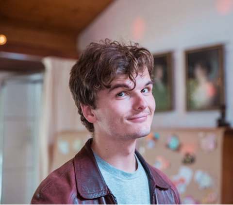 Photo of a man smiling and looking at the camera from a three-quarters angle. He has short, brown hair and is wearing a red jacket with a light blue shirt. 