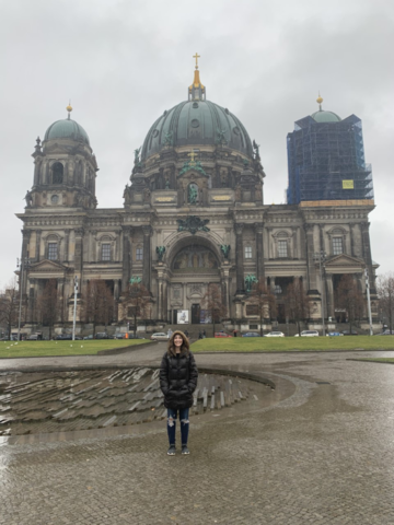 Photo of a woman in a large, black coat with a fuzzy hood standing in front of a large, ornate building. It is raining.