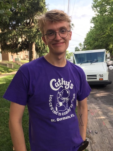 Photo of a man with blonde hair smiling at the camera. He is wearing a purple shirt and black, round glasses.