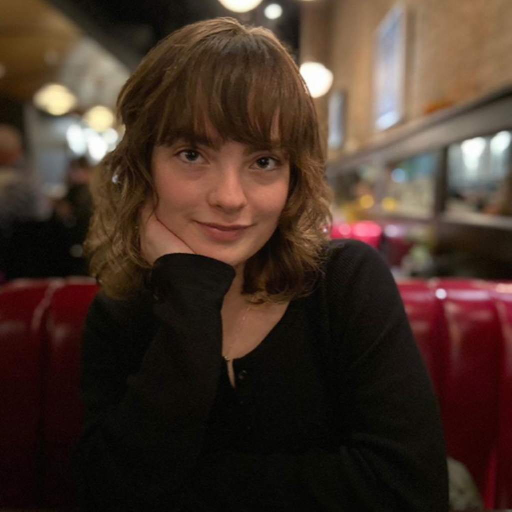 A photo of a woman with short, wavy brown hair wearing a black, long sleeved shirt. She is resting her left hand on her cheek.