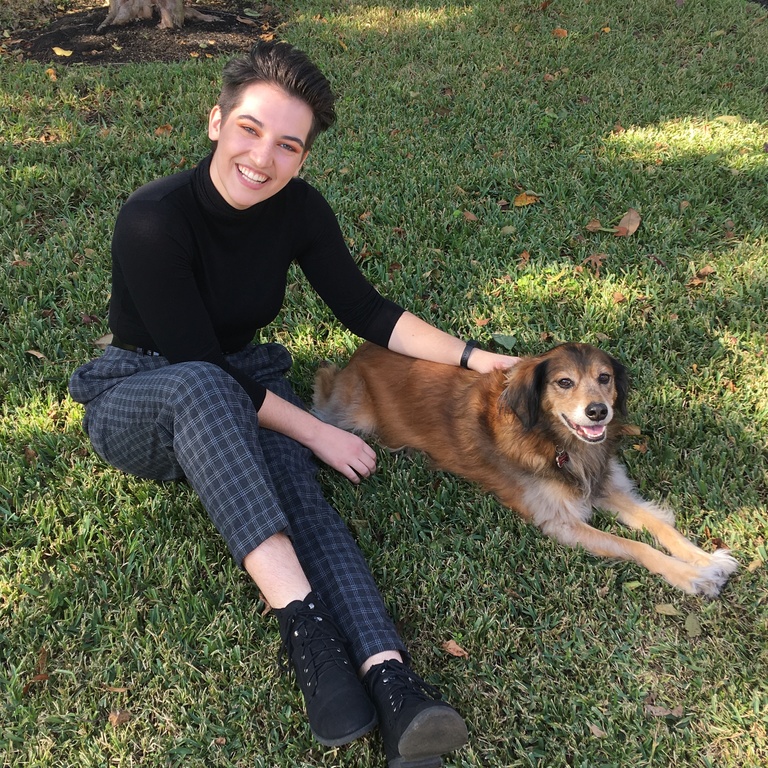 A photo of a woman with short, brown hair smiling at the camera. She is sitting on a grassy field next to a medium sized, long-furred, brown colored dog. Her right arm is petting the dog's head and her left arm is resting near the dog's foot. She is wearing a black, three-fourths length shirt, denim capris, and black boots.