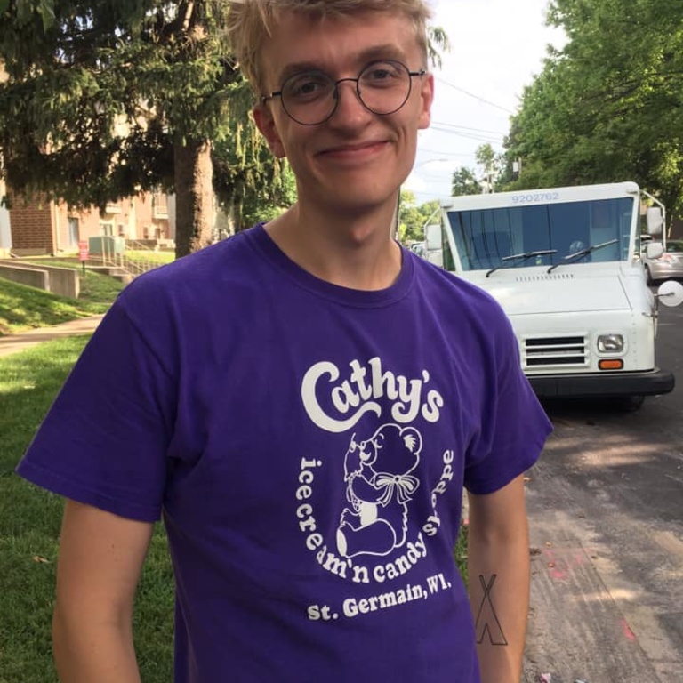 Photo of a man with blonde hair smiling at the camera. He is wearing a purple shirt and black, round glasses.